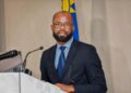 Stanley Shanapinda1 - The Brief | Namibia's Leading Business & Financial News Man in a navy suit and blue striped shirt speaks at a podium with a microphone, flag in background.