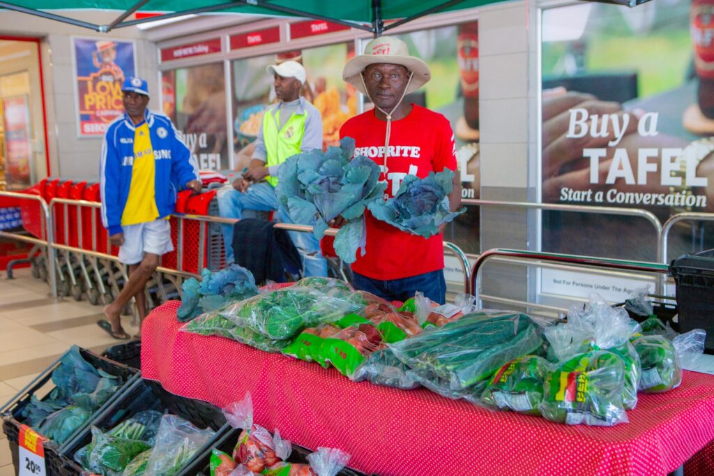 WhatsApp Image 2026-06-02 at 50544 PM - The Brief | Namibia's Leading Business & Financial News Vendor in a red shirt and wide-brim hat sells leafy greens at a market stall inside a store, with bags of vegetables on a red checkered table.