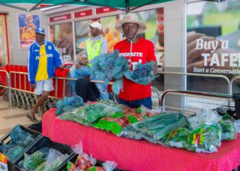 Vendor in a red shirt and wide-brim hat sells leafy greens at a market stall inside a store, with bags of vegetables on a red checkered table.