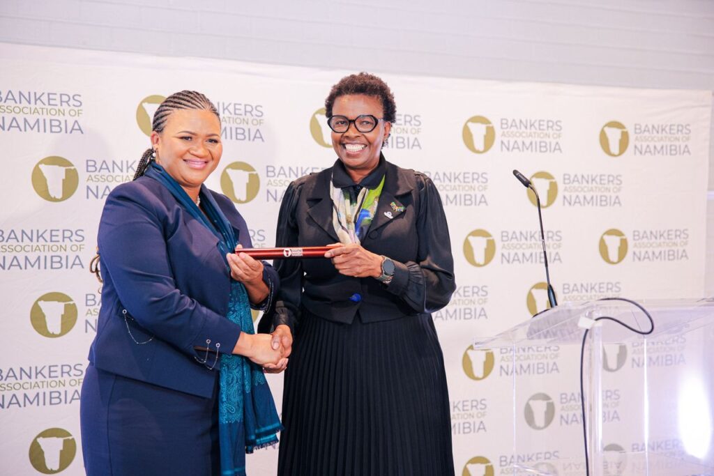WhatsApp Image 2026-06-02 at 53221 PM - The Brief | Namibia's Leading Business & Financial News Two women in formal suits shake hands on a stage, exchanging a red cylindrical award at a Bankers Association of Namibia event, with a logo backdrop behind them.