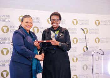 Two women in formal suits shake hands on a stage, exchanging a red cylindrical award at a Bankers Association of Namibia event, with a logo backdrop behind them.