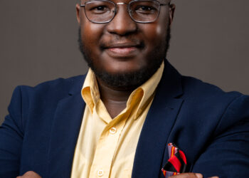 Portrait of a smiling man in a navy blazer and yellow shirt with glasses, arms crossed in front of a gray backdrop.