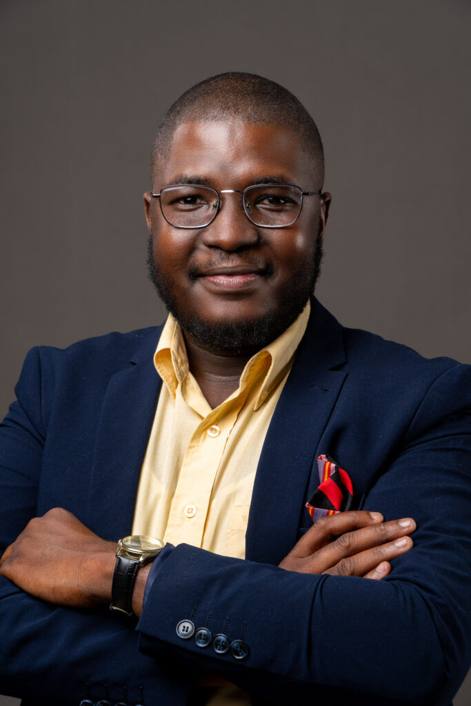 Portrait of a smiling man in a navy blazer and yellow shirt with glasses, arms crossed in front of a gray backdrop.