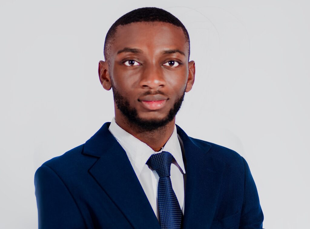 Portrait of a Black man in a navy suit and blue tie, facing the camera with a neutral expression against a light gray background