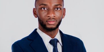 Portrait of a Black man in a navy suit and blue tie, facing the camera with a neutral expression against a light gray background