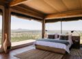Rustic bedroom with a platform bed and geometric pillows, open glass walls revealing a vast savannah landscape.