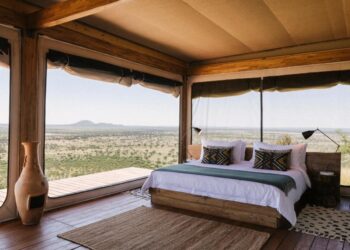Rustic bedroom with a platform bed and geometric pillows, open glass walls revealing a vast savannah landscape.