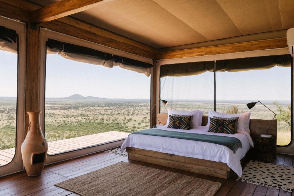 Rustic bedroom with a platform bed and geometric pillows, open glass walls revealing a vast savannah landscape.