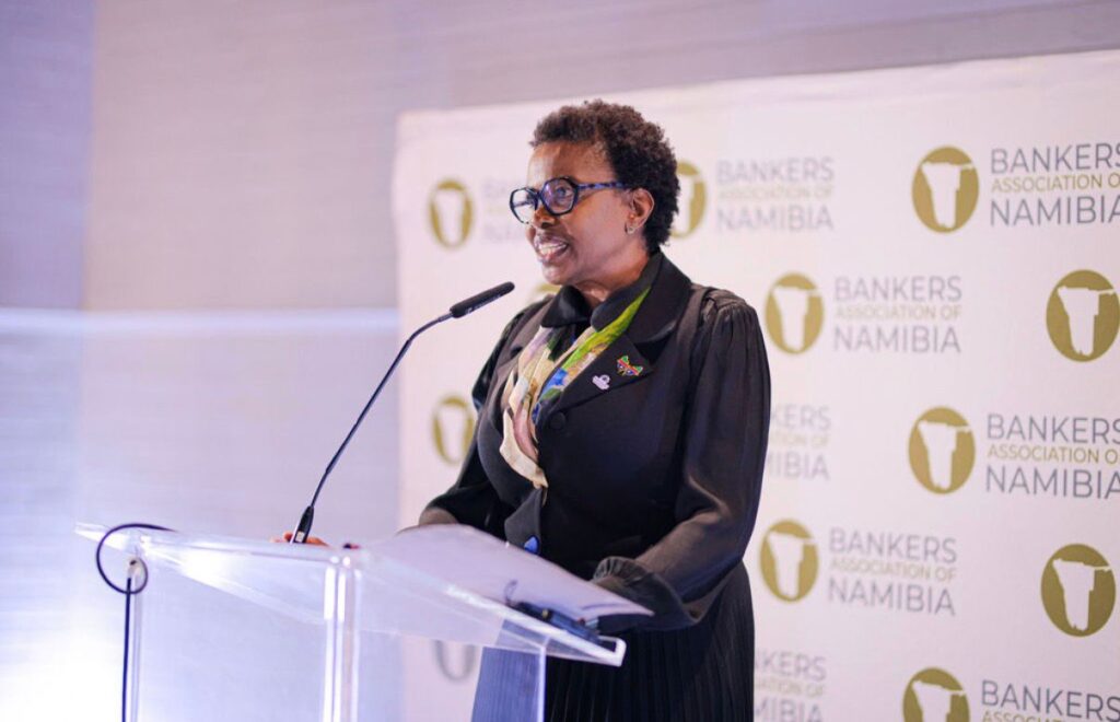 ESTER KALI3 - The Brief | Namibia's Leading Business & Financial News Black woman in a black outfit and blue glasses speaks at a clear podium, with a Bankers Association of Namibia backdrop behind her.