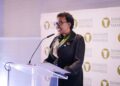 Black woman in a black outfit and blue glasses speaks at a clear podium, with a Bankers Association of Namibia backdrop behind her.