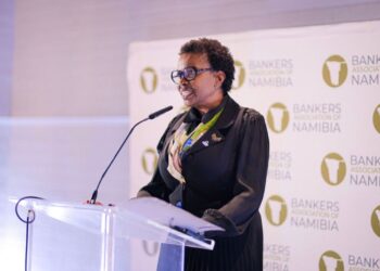 Black woman in a black outfit and blue glasses speaks at a clear podium, with a Bankers Association of Namibia backdrop behind her.