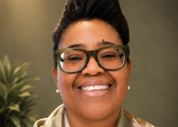 Close-up portrait of a smiling woman with large rectangular glasses, wearing a patterned scarf.
