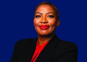 Professional headshot of a confident woman wearing a black blazer and red blouse against a blue background