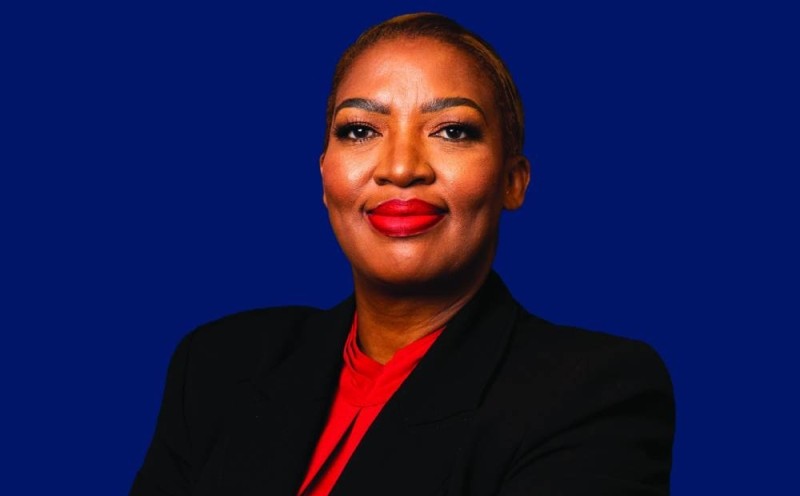 Professional headshot of a confident woman wearing a black blazer and red blouse against a blue background