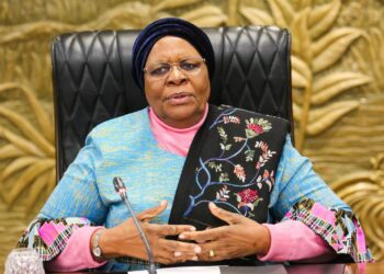 Older woman in a blue patterned jacket and pink sweater, seated at a desk with a microphone in front.