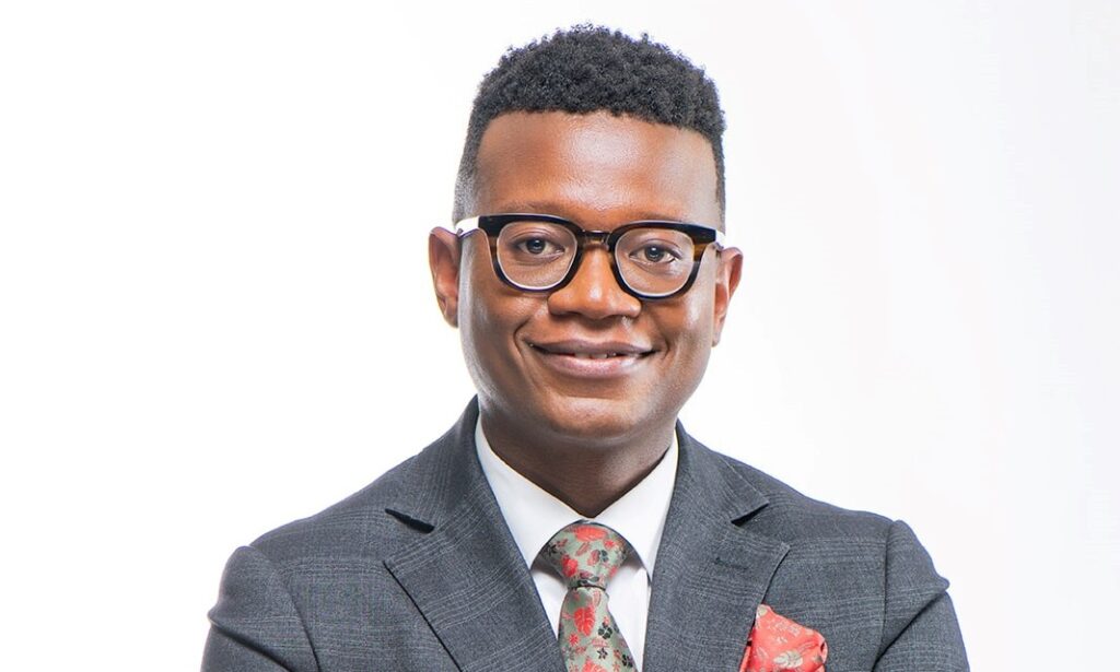 Portrait of a smiling man in a gray suit, white shirt, floral tie, and glasses against a white background.
