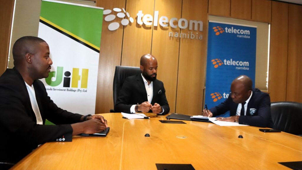 WhatsApp Image 2026-06-04 at 40444 PM - The Brief | Namibia's Leading Business & Financial News Three men in suits sit around a wooden conference table as one signs documents during a business meeting at a Telecom Namibia event area with branded banners in the background.