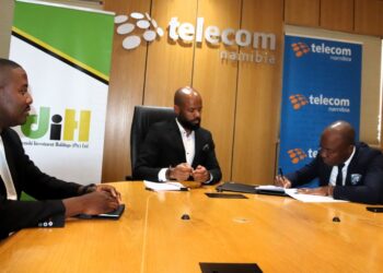 Three men in suits sit around a wooden conference table as one signs documents during a business meeting at a Telecom Namibia event area with branded banners in the background.