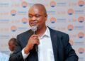 Timotheus Namwand - The Brief | Namibia's Leading Business & Financial News Man in a dark suit and white shirt speaks into a handheld microphone at a event with a branded backdrop behind him