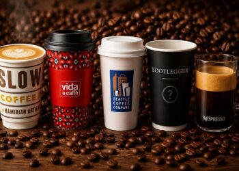 Five coffee cups in a row on a wooden surface with coffee beans scattered; from left: slow coffee cup, red patterned vida e caffè cup, white Seattle Coffee Company cup, black Bootlegger cup with a question mark, and a clear glass Nespresso mug with crema.