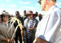 Group of people outdoors, a woman in a wide-brimmed hat and scarf speaks to a crowd while others listen in a sunny field.