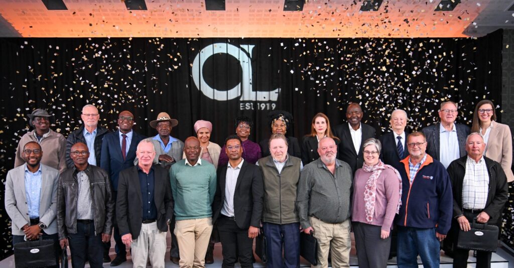 Group photo of diverse professionals on a stage with confetti, in front of a black backdrop with a large 'GL' logo and 'EST 1919' text, celebrating an anniversary.