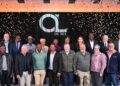 Group photo of diverse professionals on a stage with confetti, in front of a black backdrop with a large 'GL' logo and 'EST 1919' text, celebrating an anniversary.