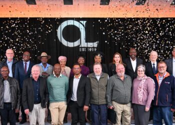 Group photo of diverse professionals on a stage with confetti, in front of a black backdrop with a large 'GL' logo and 'EST 1919' text, celebrating an anniversary.