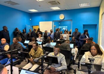 Adults in a computer lab seated at workstations while a group stands at the back of a blue-walled room.