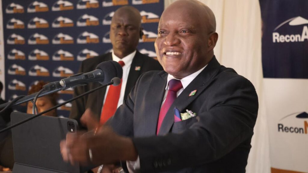 Smiling man in a dark suit and red tie speaks at a podium with microphones at a conference.