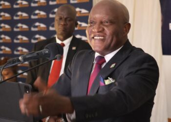 Smiling man in a dark suit and red tie speaks at a podium with microphones at a conference.
