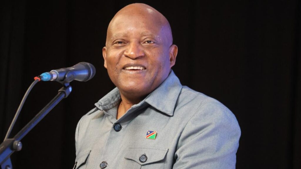 Ngurare - The Brief | Namibia's Leading Business & Financial News Smiling man at a microphone on a stage, wearing a light gray shirt with a small flag pin on the chest.