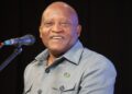 Ngurare - The Brief | Namibia's Leading Business & Financial News Smiling man at a microphone on a stage, wearing a light gray shirt with a small flag pin on the chest.