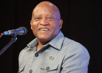 Smiling man at a microphone on a stage, wearing a light gray shirt with a small flag pin on the chest.