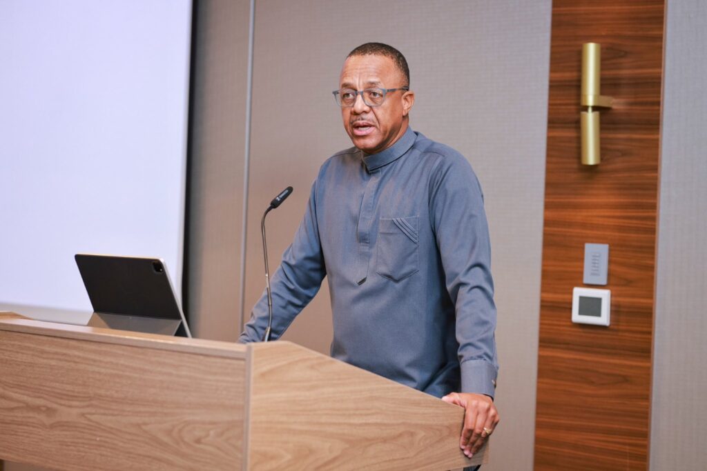 Man speaking at a wooden podium with a microphone and an open laptop in front of him, delivering a presentation in a conference room.