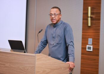 Man speaking at a wooden podium with a microphone and an open laptop in front of him, delivering a presentation in a conference room.