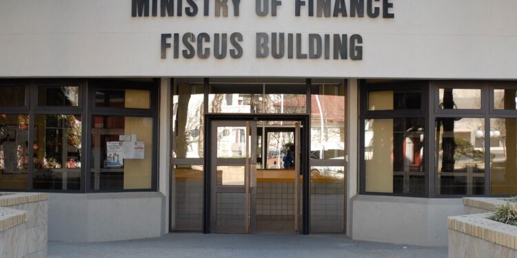 Front entrance of the Ministry of Finance, with a large sign reading 'MINISTRY OF FINANCE FISCUS BUILDING' above glass doors.