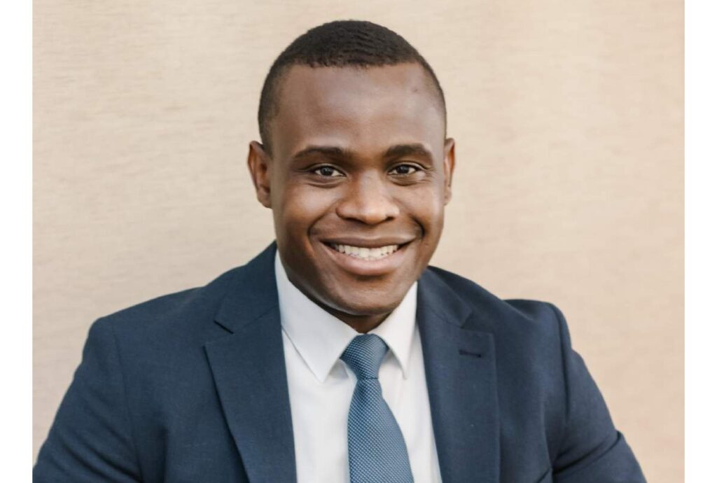 Smiling man in a navy suit and light blue tie, head-and-shoulders portrait against a beige background.