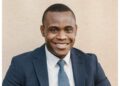 jason hailonga - The Brief | Namibia's Leading Business & Financial News Smiling man in a navy suit and light blue tie, head-and-shoulders portrait against a beige background.