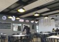 Indoor cafe scene with black chairs, white tables, and yellow pendant lights; a staff member in apron serves at the counter.