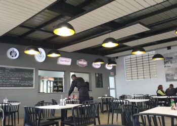 Indoor cafe scene with black chairs, white tables, and yellow pendant lights; a staff member in apron serves at the counter.