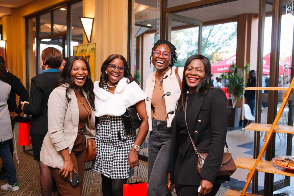 WhatsApp Image 2026-06-09 at 33214 PM - The Brief | Namibia's Leading Business & Financial News Four women smiling and posing for a photo at an indoor event, each holding a red gift bag or shopping bag, dressed stylishly.