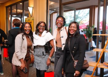 Four women smiling and posing for a photo at an indoor event, each holding a red gift bag or shopping bag, dressed stylishly.