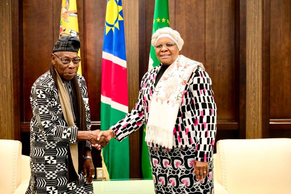 Two formally dressed officials shake hands in front of national flags in a wooden-paneled room.