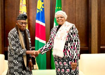 Two formally dressed officials shake hands in front of national flags in a wooden-paneled room.