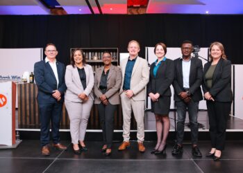 Group of seven professionals in business attire posing on a stage for a photo at a corporate event, smiling at the camera.