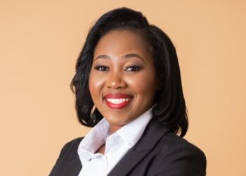 Portrait of a smiling professional woman in a black blazer and white shirt against a beige background.
