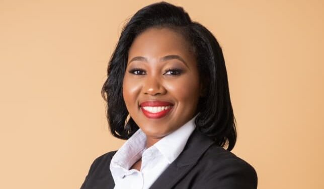 Portrait of a smiling professional woman in a black blazer and white shirt against a beige background.