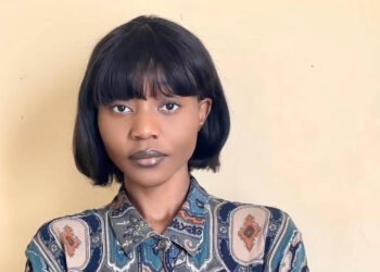 Portrait of a young woman with a short black bob and bangs, wearing a blue‑beige patterned shirt, standing against a pale yellow wall.