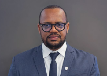 Head-and-shoulders portrait of a man in a navy suit and glasses, looking at the camera, with a gray background.